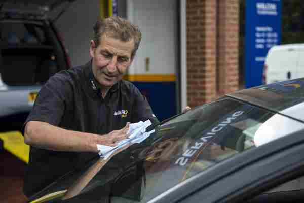 A Man in a Ash Road Service top wiping a windscreen of a car during an MOT test